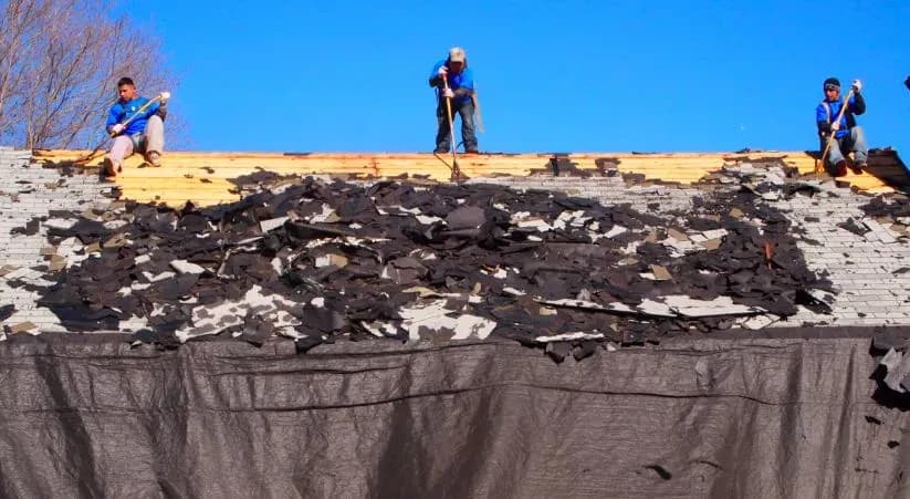 Three workers in blue shirts remove old shingles from a roof under a clear blue sky, tools in hand and debris scattered below, conveying teamwork.