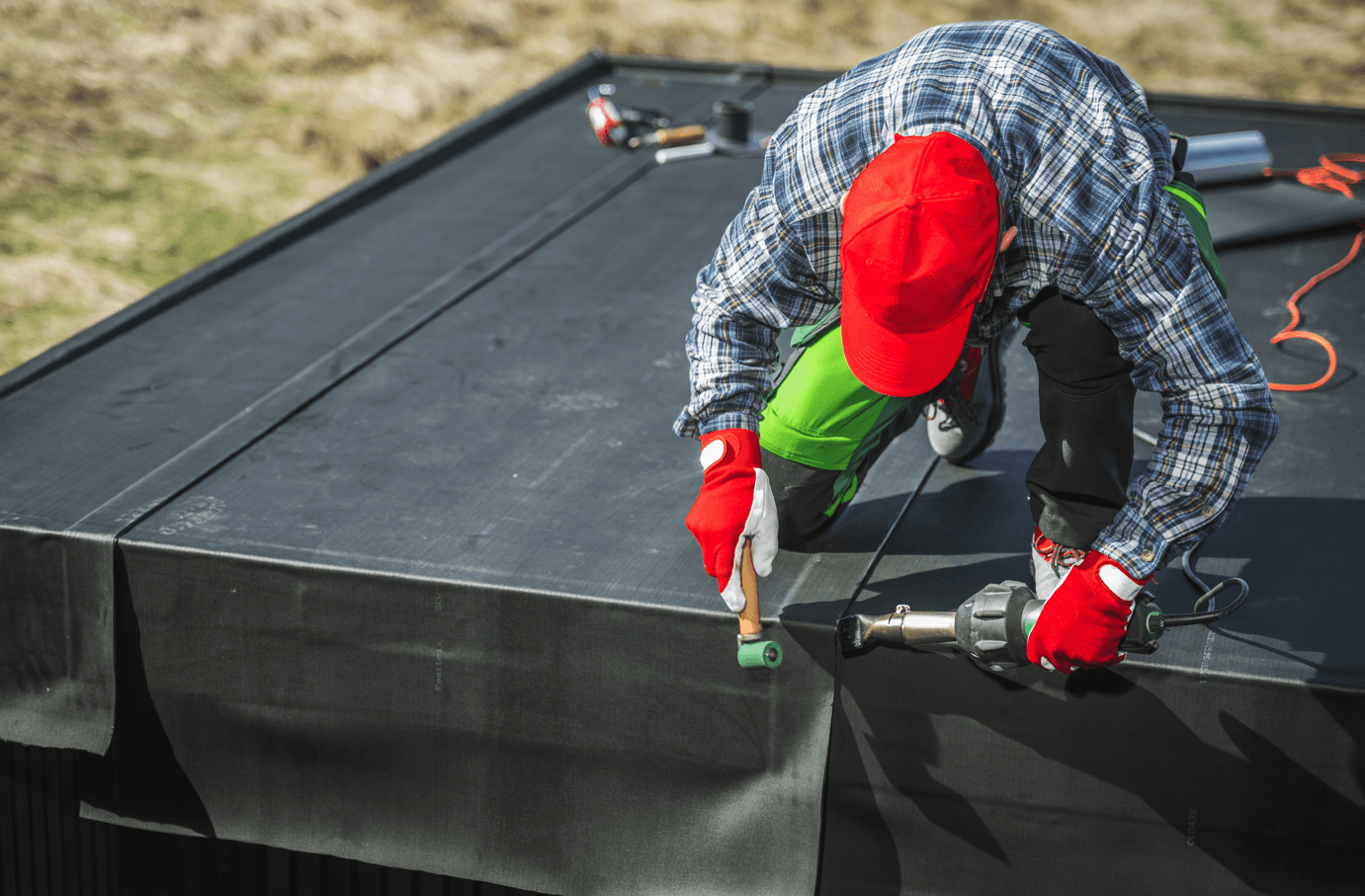 A worker in a red cap and gloves installs a black rubber roof membrane. They use a heat gun and roller, showing focus and skilled labor.