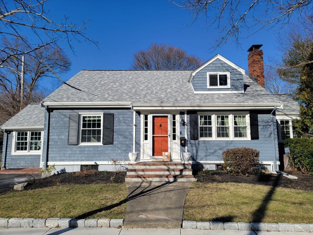 A charming blue house with a red front door, black shutters, and a brick chimney. A stone path leads to the entrance, surrounded by a neat lawn.