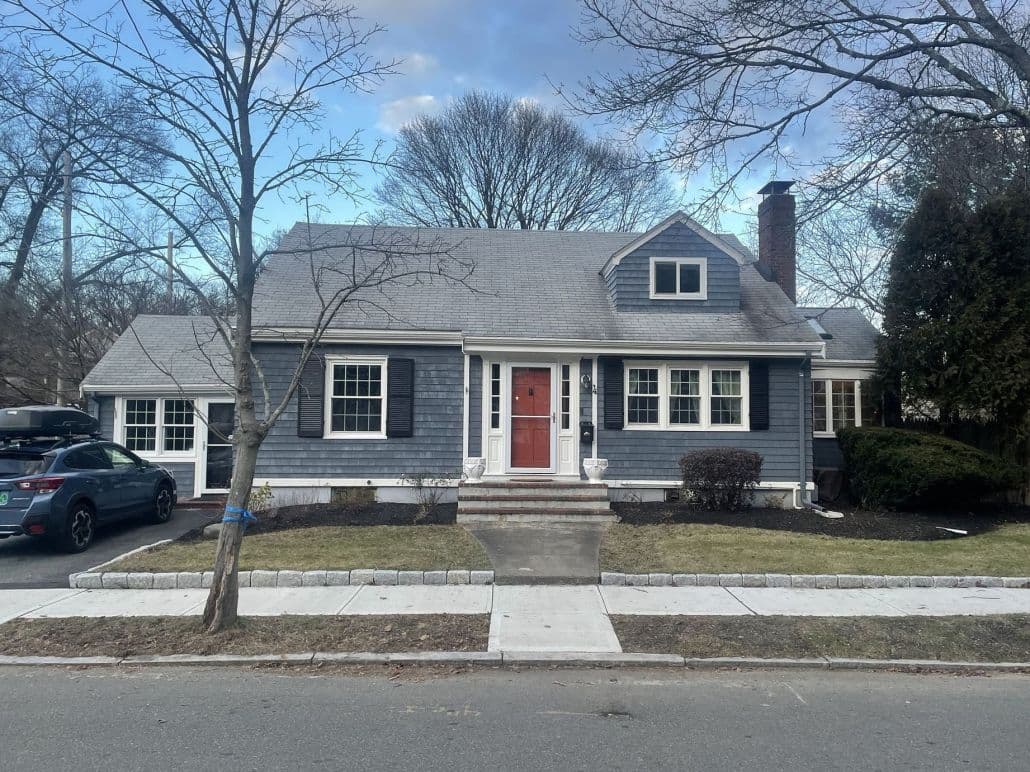 A charming blue house with a gray roof and a red door, framed by bare trees. A car is parked in the driveway. The scene feels calm and inviting.