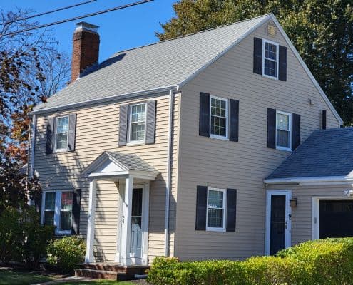 A beige colonial-style house with gray shutters and a steep roof, featuring a prominent white front door, chimney, and well-kept shrubs, under a clear blue sky.