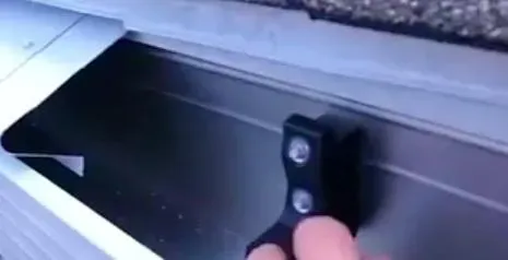 A hand pushes a black button with two screws inside a metallic gutter system under a roof edge. The scene suggests maintenance or installation work.