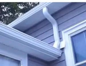A gray house exterior with white trim and a white gutter awkwardly bending around a corner. Trees are visible in the background under a cloudy sky.