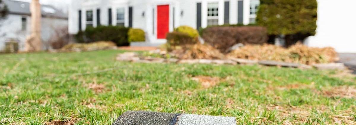 Close-up of a small gravestone on a grassy lawn with a blurred white house in the background. The house has black shutters and a striking red door.