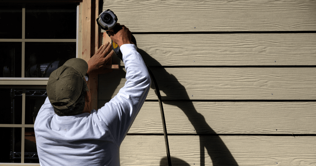 A person wearing a cap uses an electric drill on wooden siding near a window. The scene is sunlit, casting a strong shadow on the siding.
