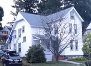 White two-story house with dormer windows and gray roof, surrounded by trees. A bare tree stands in front, and a car is parked nearby. Calm suburban setting.