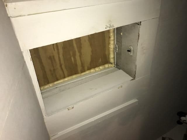 A partially open attic hatch with white wooden panels and a visible plywood interior. Foam insulation is seen around the edges, giving a construction feel.