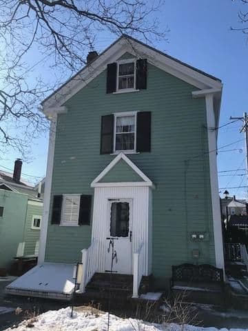 A small, light green house with black shutters and a white door and trim, under a clear blue sky. Snow covers the ground, creating a chilly, serene atmosphere.