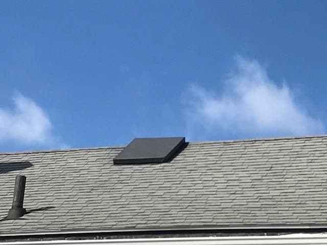 A gray, shingled roof under a clear blue sky with scattered clouds. Features a small square skylight and a vent, evoking a calm, sunny day.