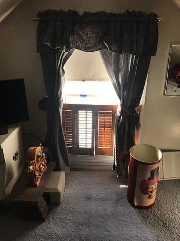 A small, sunlit attic window with gray curtains and wooden shutters. Nearby, a vintage clock and a tall cylindrical tin add a cozy, nostalgic feel.