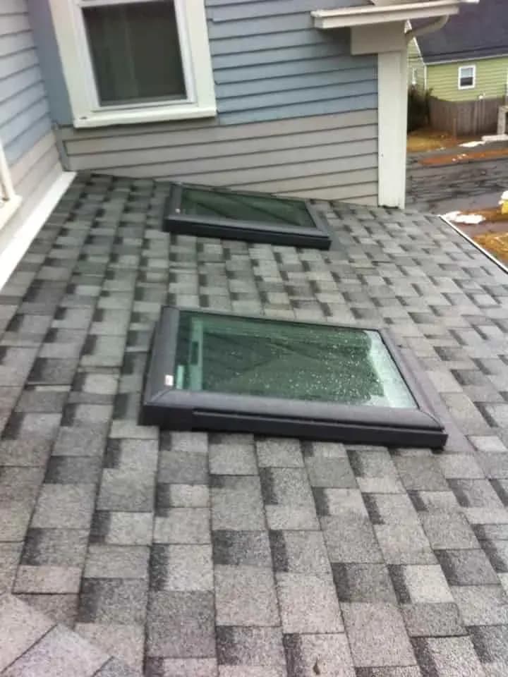 The image shows two skylights installed on a sloped gray shingle roof of a house. The sky is overcast, and the mood is calm and slightly somber.