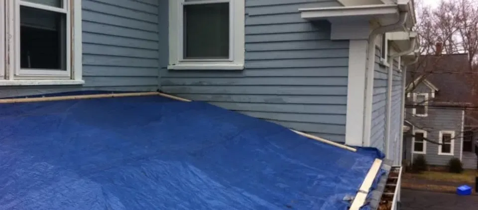 A flat roof covered with a blue tarp is attached to a light blue house. Two windows overlook the tarp, set against the backdrop of bare trees and another house.