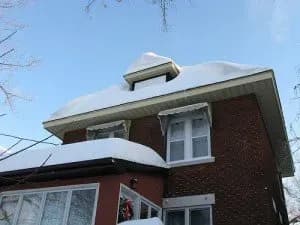 A brick house topped with a thick layer of snow under a clear blue sky. The roof has a dormer window, adding to the wintry, serene atmosphere.