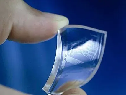A close-up of fingers holding a flexible, transparent electronic circuit. The background is a solid blue, highlighting the advanced technology.