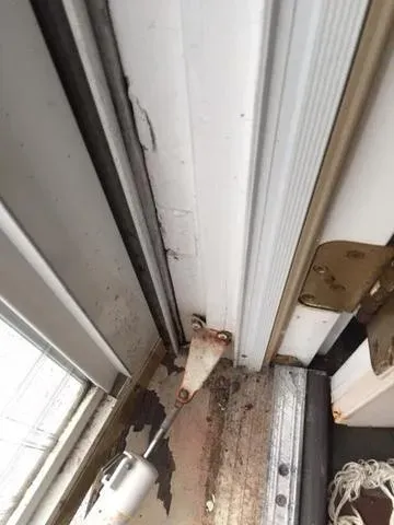 Damaged door frame with peeling paint and rusted hinge at the bottom, surrounded by chipped wood and dirt, creating a neglected and worn appearance.