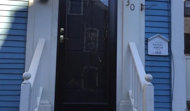 Black door with a glass panel centered on a historic blue wood-paneled house, flanked by white railings. A plaque states, "Built for Daniel Abbott, AD 1812."