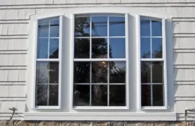 A white, three-panel bay window with grid-patterned glass set in a light-colored shingled wall, reflecting trees and a soft, sunny day.