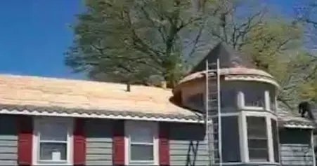 A house with gray siding and red shutters under a clear blue sky. A ladder leans against a turret, and a man is working on the roof.