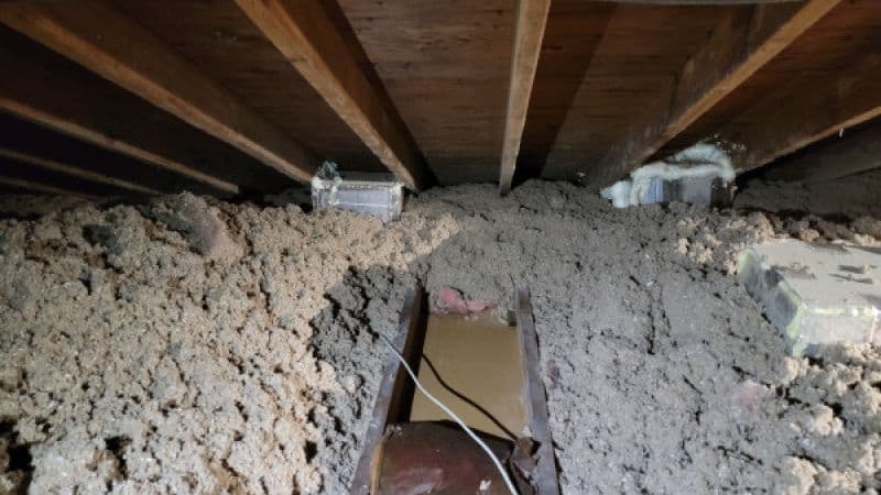 An attic space with exposed wooden beams and insulation scattered around. Loose, dusty debris covers the floor, with a few electrical wires visible.