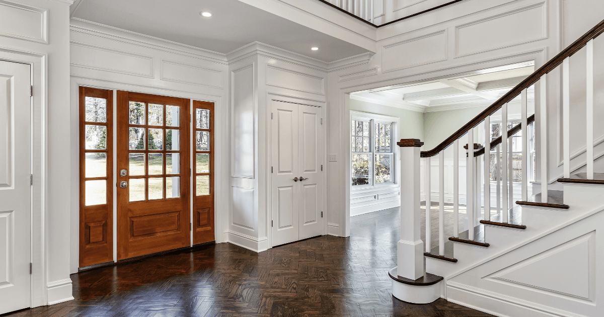 Elegant foyer featuring wooden double doors with glass panels, white paneled walls, dark herringbone flooring, and a curved staircase leading upstairs.