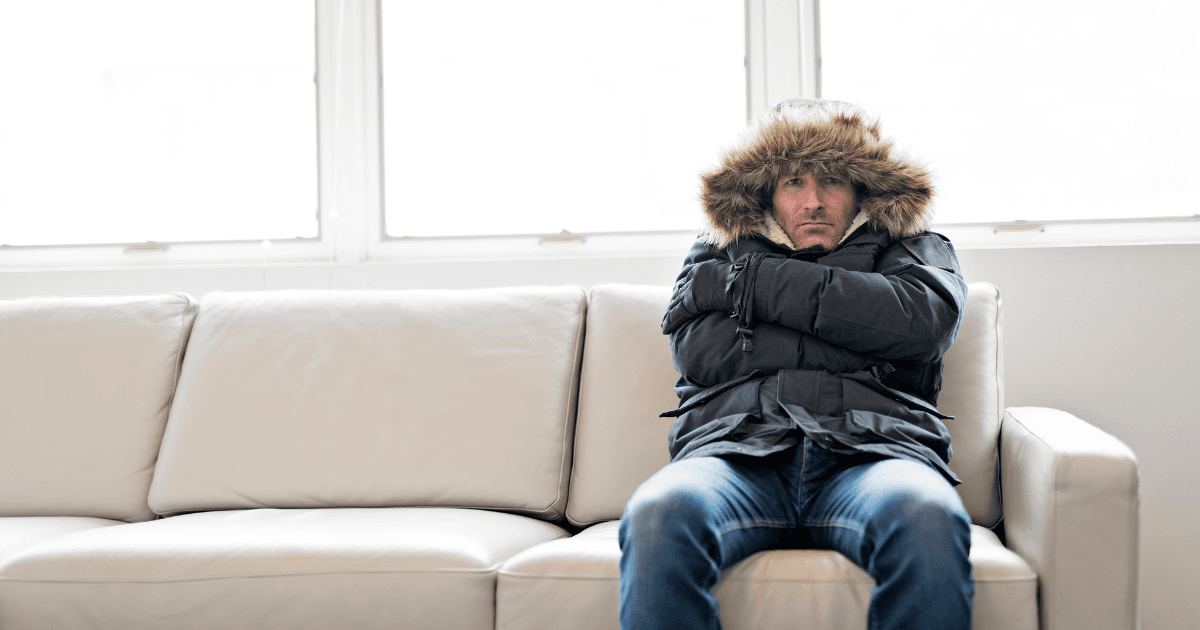 A person in a parka with a fur-lined hood sits huddled on a white sofa, arms crossed, conveying a sense of cold or discomfort, in a brightly lit room.