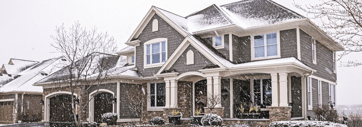 Two-story house with snow-covered roof and trees, featuring stone and gray siding. Snowflakes gently fall, creating a serene winter scene.