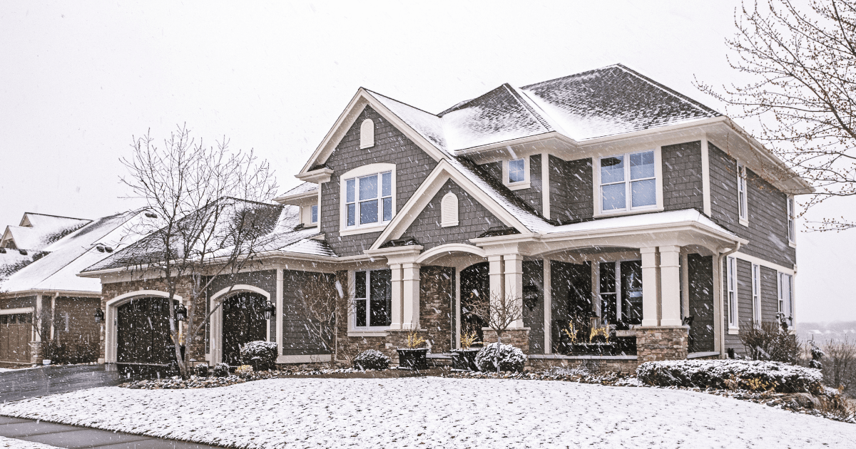 A two-story house with a snow-dusted roof sits amid a gentle snowfall. The modern architecture features large windows and stone accents, creating a cozy winter scene.