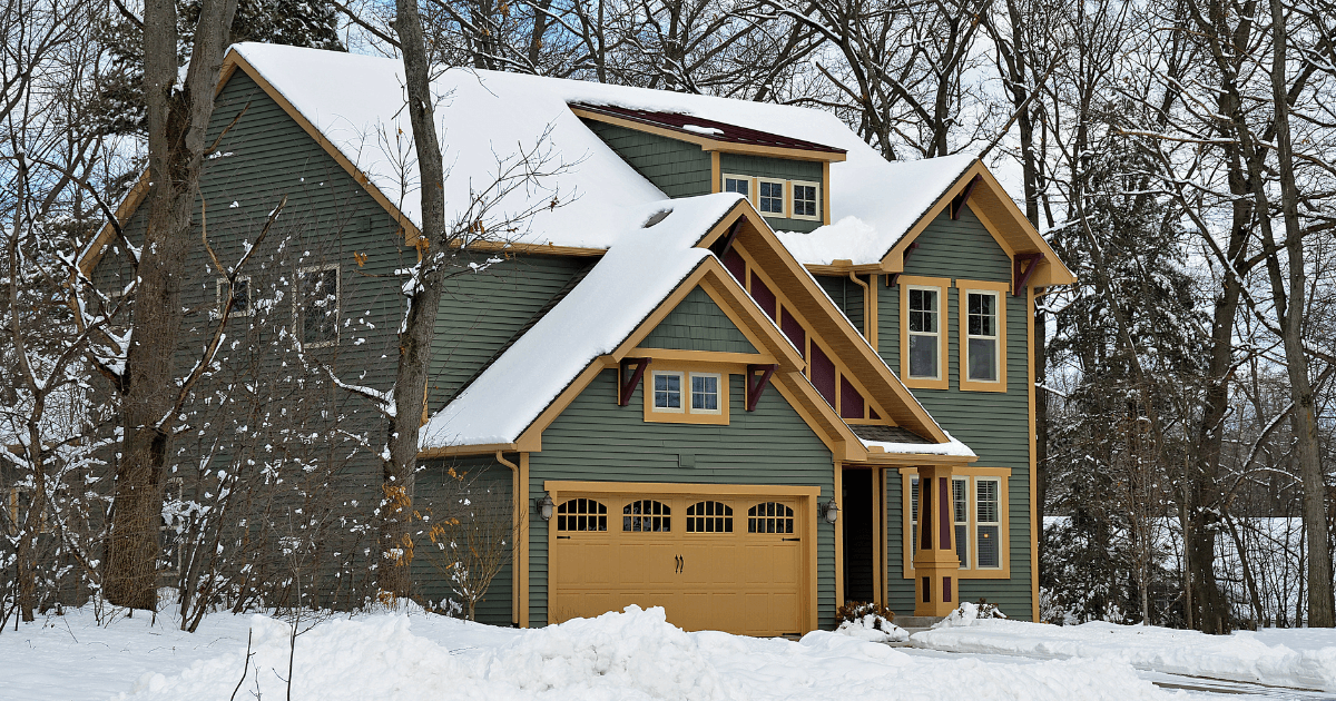 A charming two-story house with green siding and mustard trim is surrounded by snow-laden trees. The scene is peaceful and wintry.