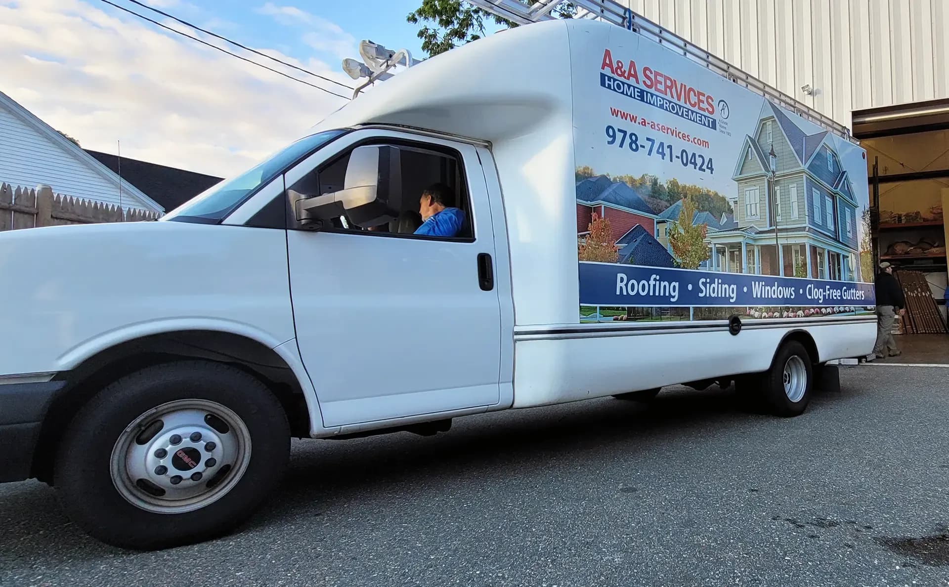 A white service van with a large side advertisement for A&A Services Home Improvement, featuring roofing, siding, and windows. A person is seated inside.