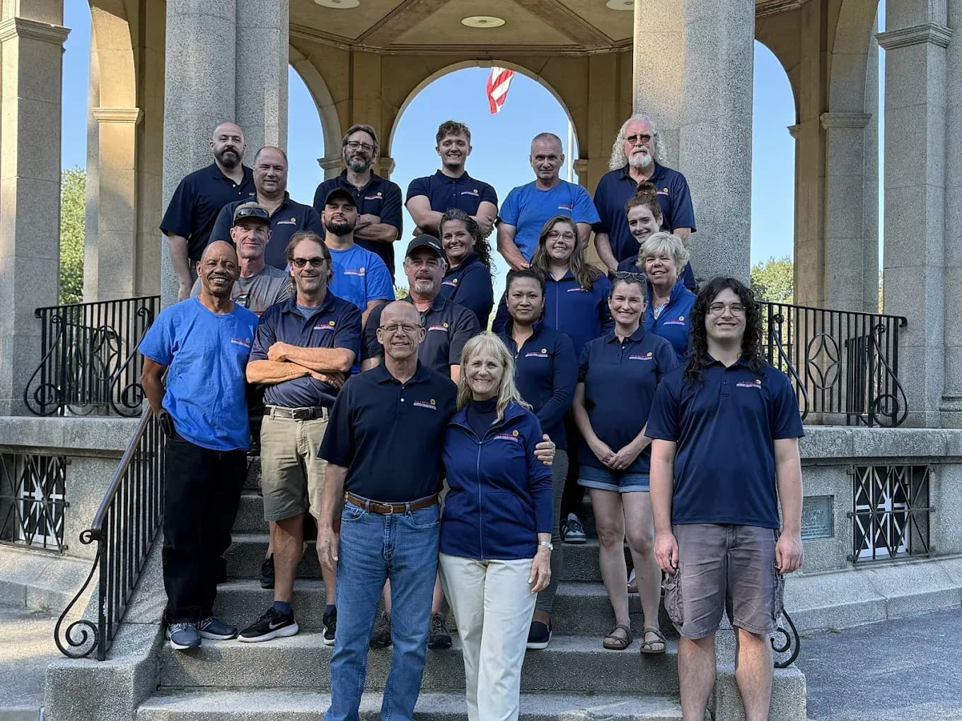 A diverse group of 19 people standing on stone steps in front of a gazebo. Most wear matching blue shirts, conveying a sense of unity and collaboration.