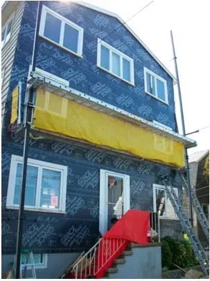 A two-story house under renovation, covered in blue building wrap. There's a yellow protective netting on the upper floor and a red carpet on the stairs.