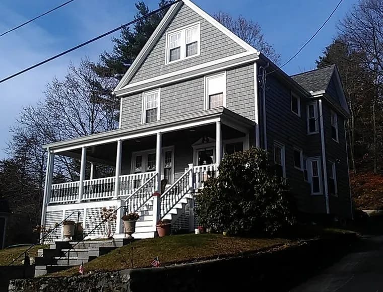 A charming gray two-story house with a steep roof and white-trimmed porch in a residential area. Surrounded by trees, it sits atop a small hill.