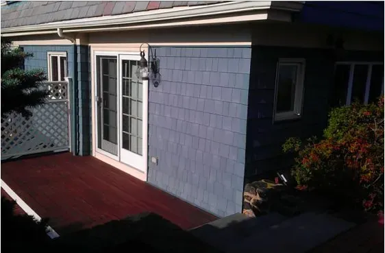 Sunny deck attached to a blue shingled house, featuring French doors, a wall-mounted lantern, and partial view of a garden with green foliage.