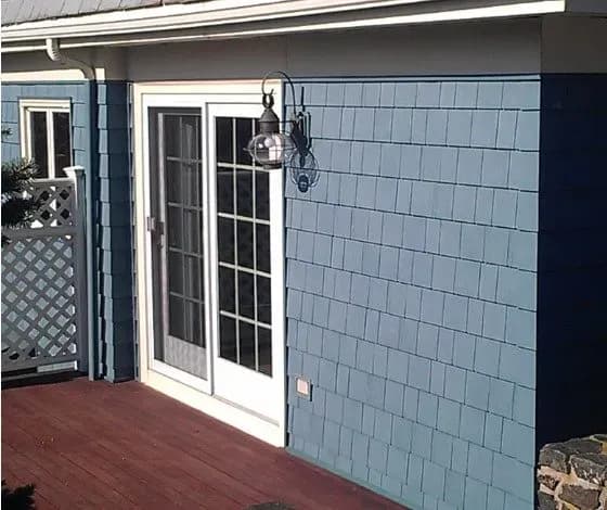 A white-framed glass patio door is set in a blue shingled wall. A black lantern with a glass globe hangs nearby, casting a gentle shadow.