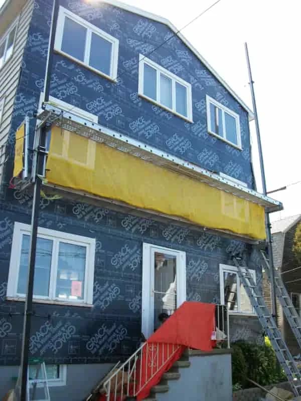 Three-story house under construction, wrapped in blue Grace building paper. A yellow tarp and red ladder lead to the entrance, suggesting ongoing work.
