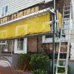 A house exterior under renovation with a yellow tarp covering exposed wall sections. Ladder and greenery nearby, conveying an active construction scene.