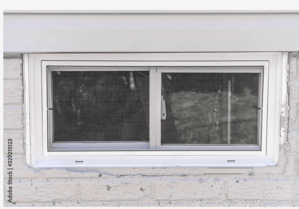A rectangular basement window with a white frame and mesh screen. It is set in a brick wall, with faint reflections visible on the glass panels.