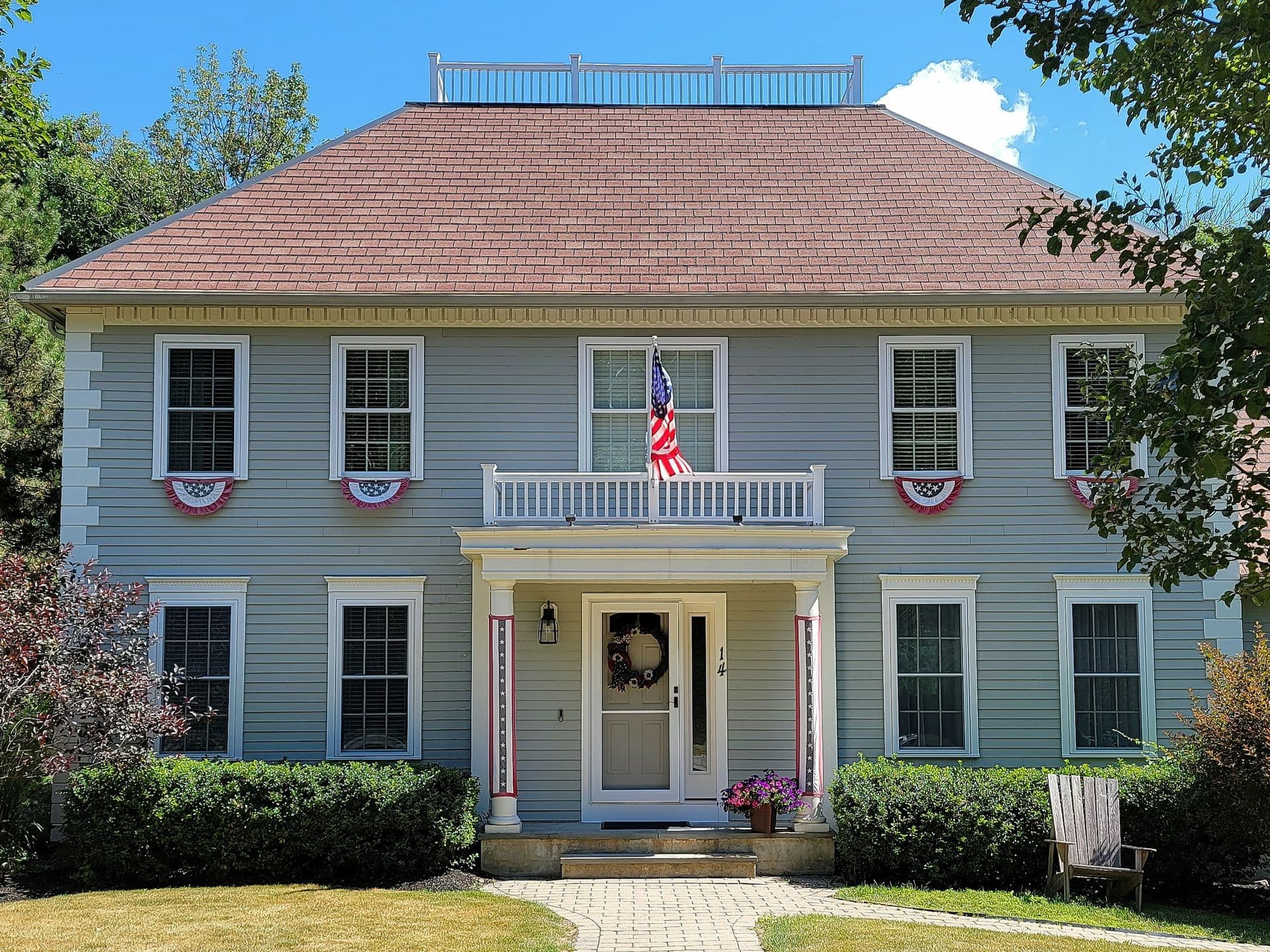 Classic two-story house with a red roof, gray siding, American flags, and patriotic banners. Trimmed hedges surround the front path, conveying a festive, welcoming atmosphere.