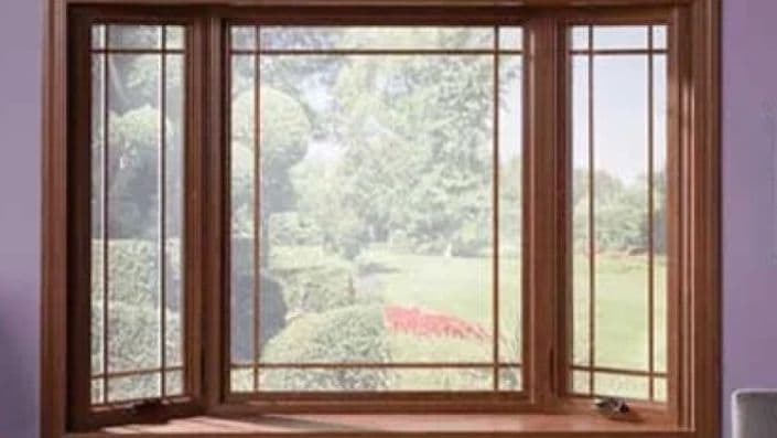Wooden-framed bay window showing a lush green garden with neatly trimmed hedges and trees under a clear sky, creating a serene, inviting view.