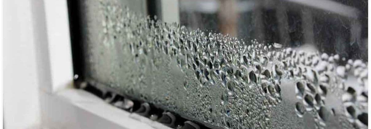 Close-up of a window with condensation forming droplets on the glass, creating a textured pattern. The scene conveys a cool, damp atmosphere.