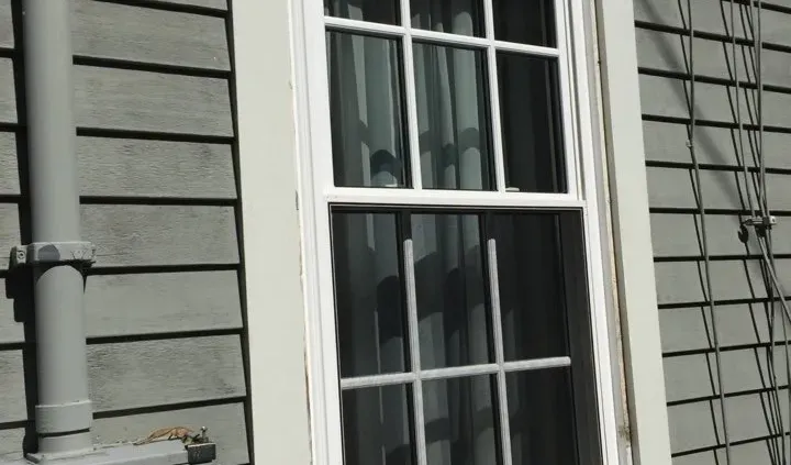 A white-framed window with grid panes is set in a gray, horizontally paneled wall. A vertical pipe runs alongside, casting shadows in the sunlight.