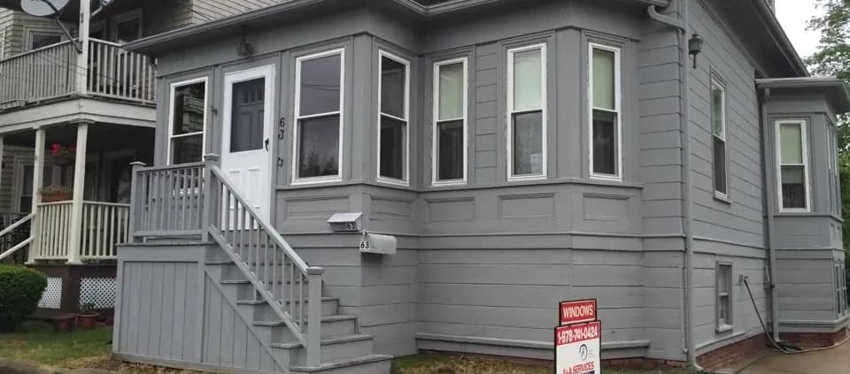A gray, two-story house with white trim and multiple windows, featuring a small front porch with stairs. A "Windows for Sale" sign is on the lawn.