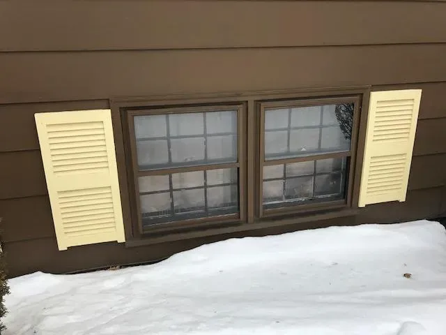 Brown house with a low-set window, partially covered by snow, has yellow shutters awkwardly placed next to it, creating a humorous mismatch.