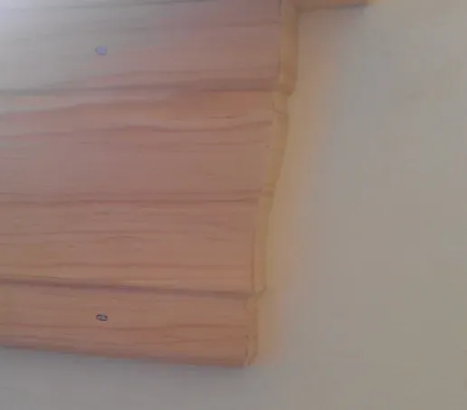 Close-up of a wooden wall panel with uneven edges, attached to a light-colored wall. The scene has a rustic feel with visible wood grain and nails.
