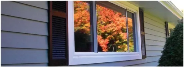 A double-pane window reflects vibrant autumn foliage, featuring red and orange leaves. The house siding is beige with dark shutters, creating a calm and cozy feel.