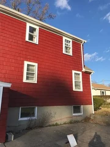Red-sided house with white trim, featuring five windows. A shadow is cast on the side, with a clear blue sky above, creating a serene suburban setting.