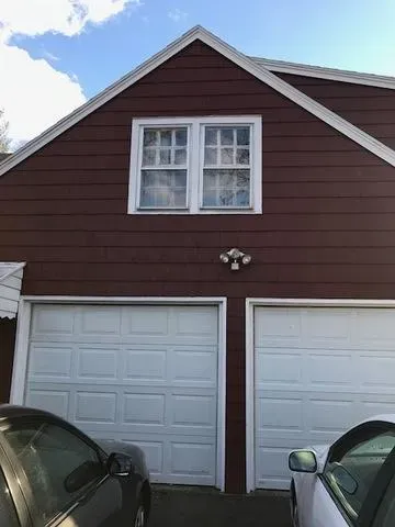 A brown house with white trim features two garage doors side by side. A double window is above the doors. Parked cars are partially visible in front.