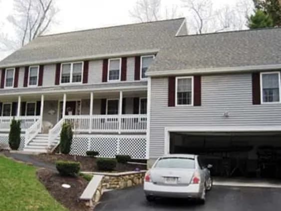 Two-story suburban house with gray siding, white trim, and burgundy shutters. A white car is parked in the driveway next to an open garage. Calm scene.