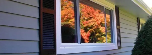A large window reflects vibrant autumn trees with orange and red leaves. It's set in a house's grey siding, flanked by dark shutters, evoking a cozy, warm atmosphere.